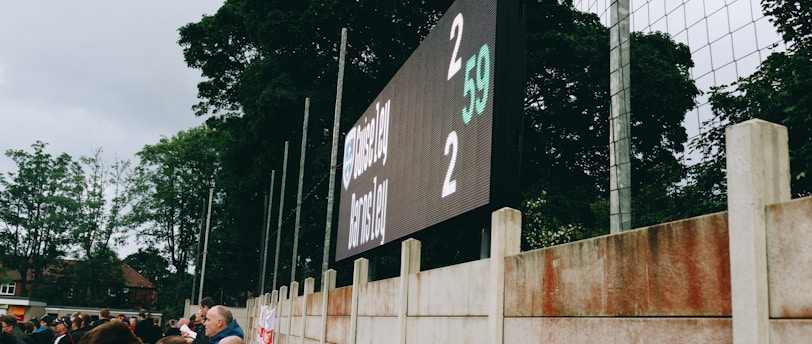 A large digital scoreboard at an outdoor sports venue displays a score of 2-2. Spectators are gathered, observing the event with interest. The scene is enclosed by a tall metal fence and surrounded by lush green trees, indicating a natural setting. Overcast skies add a muted tone to the environment.