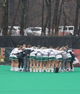 A group of athletes huddled together in a circle on a green sports field. They are wearing uniforms with green shorts and white tops, indicating a team sport environment. Lacrosse sticks are lined up against a fence in the background. Trees and parked cars are visible beyond the fence, suggesting an outdoor location.