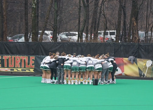 A group of athletes huddled together in a circle on a green sports field. They are wearing uniforms with green shorts and white tops, indicating a team sport environment. Lacrosse sticks are lined up against a fence in the background. Trees and parked cars are visible beyond the fence, suggesting an outdoor location.