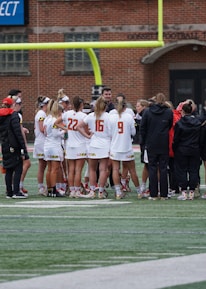 A group of athletes wearing sports uniforms huddles on a grassy field, likely preparing or strategizing for a game. The players are surrounded by additional team members or coaches wearing black jackets. A goalpost and net are visible in the background, along with a brick building.