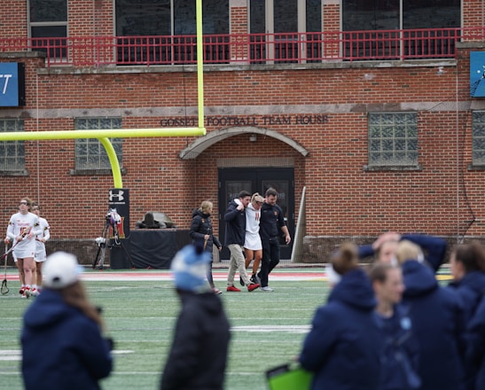 A group of people on a sports field, with some dressed in athletic gear, and others in casual clothing. A brick building labeled 'Gosset Football Team House' is in the background. One of the athletes appears to be aided by others, suggesting a possible injury.