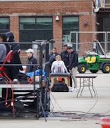 A group of people are gathered around an individual wearing a sports jersey with the number 11. They appear to be in a sports facility area, possibly a training or medical setup. There are tables, chairs, and equipment scattered around, with a brick building in the background. Some individuals are seated on a platform with electronics close by, suggesting a recording or monitoring activity. Utility vehicles and an orange barrier are present in the background.
