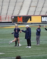Several people are on a sports field with an empty bleacher in the background. One person is high-fiving a coach, while others stand nearby. They are wearing athletic clothing, hinting at a team sport environment. A sign with the text 'Muscle Milk' is visible in the background.