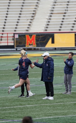 Several people are on a sports field with an empty bleacher in the background. One person is high-fiving a coach, while others stand nearby. They are wearing athletic clothing, hinting at a team sport environment. A sign with the text 'Muscle Milk' is visible in the background.