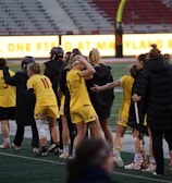 A group of athletes wearing yellow sports uniforms are standing on a sports field. Some are holding lacrosse sticks, and they appear to be interacting casually. Several people are dressed in black coats. In the background, there is a bright sign with text and parts of a stadium are visible.