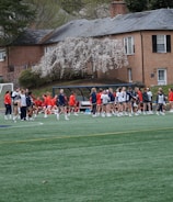 A group of people, primarily young athletes, are gathered on a sports field, some dressed in uniforms consisting of navy blue and red colors. In the background, there are several red-brick buildings and blossoming trees, suggesting a springtime setting. A goalpost can be seen, indicating that this is likely a game of soccer or lacrosse. The scene is lively and bustling with activity.