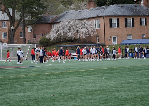 A group of people, primarily young athletes, are gathered on a sports field, some dressed in uniforms consisting of navy blue and red colors. In the background, there are several red-brick buildings and blossoming trees, suggesting a springtime setting. A goalpost can be seen, indicating that this is likely a game of soccer or lacrosse. The scene is lively and bustling with activity.