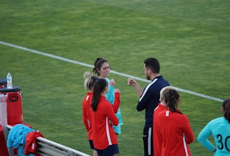 A group of people wearing sports attire are gathered on a grassy field. Some individuals are dressed in red jackets, while others are in turquoise and navy blue. A coach appears to be giving instructions. Sports equipment, such as water bottles and a bench with towels, can be seen nearby.