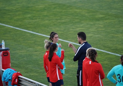 A group of people wearing sports attire are gathered on a grassy field. Some individuals are dressed in red jackets, while others are in turquoise and navy blue. A coach appears to be giving instructions. Sports equipment, such as water bottles and a bench with towels, can be seen nearby.