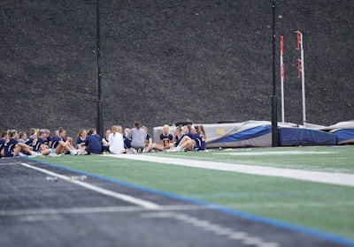 A group of people, likely a sports team, is sitting together on a field. They are wearing matching athletic uniforms, suggesting team unity, and seem to be engaged in a discussion or meeting. In the background, there is sports equipment and a high fence.