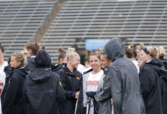 A group of people, likely athletes, are gathered together on a sports field. They are wearing black jackets with some individuals dressed in white sports uniforms underneath. A few people have their hair tied up in buns, and most are engaged in conversation. The background shows tiered stadium seats, suggesting the setting is an outdoor sports venue.