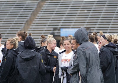 A group of people, likely athletes, are gathered together on a sports field. They are wearing black jackets with some individuals dressed in white sports uniforms underneath. A few people have their hair tied up in buns, and most are engaged in conversation. The background shows tiered stadium seats, suggesting the setting is an outdoor sports venue.