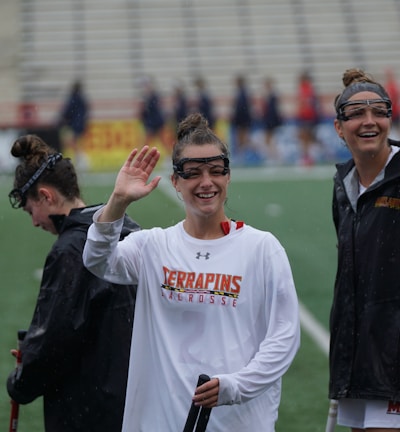 Several athletes in sports attire are on an outdoor sports field, interacting and smiling. One individual in the foreground waves with a friendly expression, while another stands beside her, also smiling. They wear protective goggles and gear associated with lacrosse. The background features indistinct figures in dark clothing against a backdrop of stadium seating.
