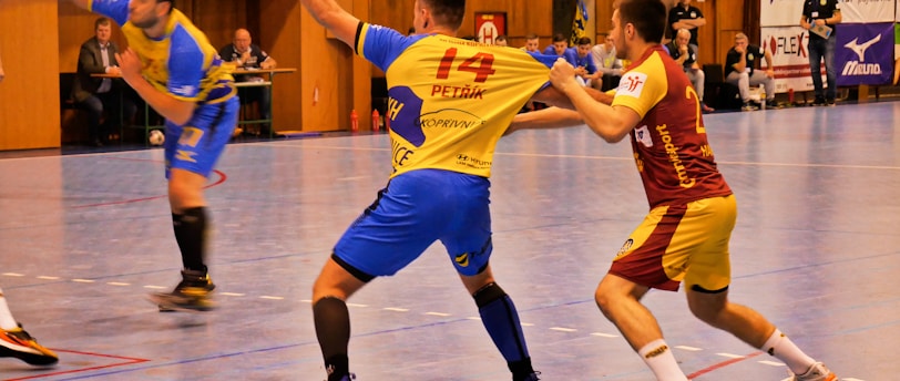 A dynamic indoor sports scene with players in motion on a court. Two athletes in blue and yellow uniforms are engaged in an intense handball game, with one player gripping the other's jersey. The background shows other players, several seated people, and advertisements on the walls.