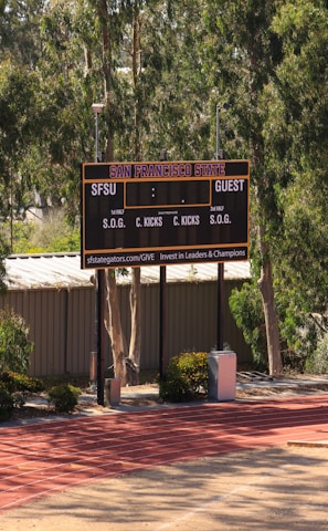 An outdoor scoreboard for San Francisco State is seen among trees and greenery. The board displays sections for shots on goal and corner kicks, with areas for both home and guest teams. It is situated near a running track with a building in the background.